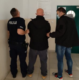 Policjanci trzymają z dwóch stron zatrzymanego mężczyznę. Znajdują się w korytarzu, przed wejściem do aresztu.