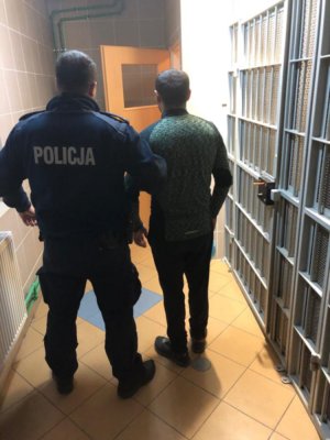 Umundurowany policjant przeprowadza przez korytarz zatrzymanego mężczyznę w ciemnej kurtce.