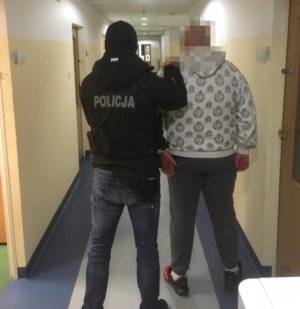 Policjant trzyma zatrzymanego mężczyznę, ubranego w szarą bluzę na korytarzu w komendzie policji