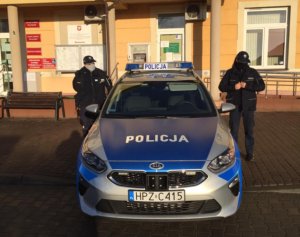 Oznakowany policyjny radiowóz zaparkowany przed budynkiem urzędu gminy w Prażmowie. Po obu stronach samochodu stoją dwie umundurowane policjantki