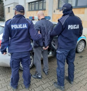 Umundurowani policjanci doprowadzają mężczyznę w ciemnej kurtce do oznakowanego radiowozu