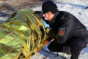 Umundurowany policjant prezentuje nałożenie foli izotermicznej na osobę wymarzniętą. Złotym kolorem na wierzchu