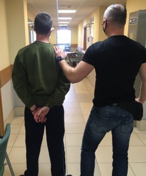 Nieumundurowany policjant z lewej strony znajduje się na korytarzu sadowym z zatrzymanym mężczyzną w zielonej bluzie