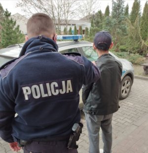 Umundurowany policjant stoi przed radiowozem wraz z zatrzymanym mężczyzną w ciemnej kurtce