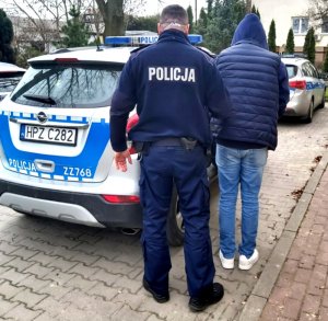 Umundurowany policjant prowadzi zatrzymanego mężczyznę w granatowej kurtce z kapturem. Obok znajdują się dwa policyjne radiowozy