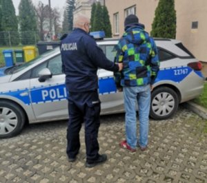 Umundurowany policjant wraz zatrzymanym w kolorowej kurtce przed radiowozem