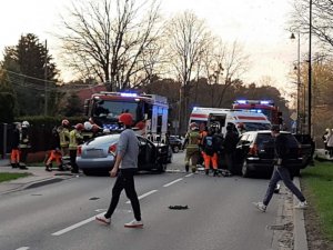 Na jezdni znajdują się dwa rozbite samochody, strażacy ich pojazd oraz przechodnie