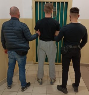 Młody mężczyzna pomiędzy nieumundurowanymi policjantami przed pomieszczeniem aresztu.