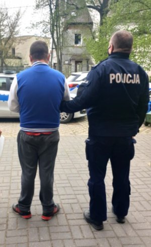 Umundurowany policjant prowadzi zatrzymanego. W tle policyjny radiowóz.