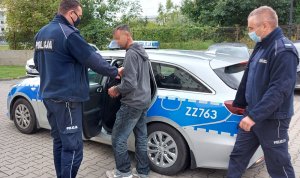 Policjanci z zatrzymanym mężczyzną przed radiowozem na dziedzińcu komendy