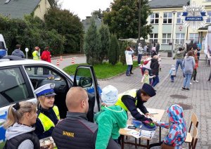Policjantka wydziału ruchu drogowego rozmawiająca z uczestnikami pikniku, obok dzieci kolorujące rysunki