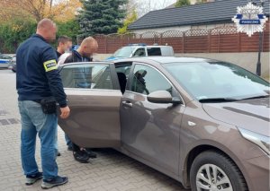 Zatrzymany mężczyzna z policjantami na dziedzińcu komendy wsiadający do radiowozu nieoznakowanego