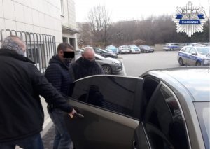 Nieumundurowani policjanci wraz z zatrzymanym mężczyzną przed wejściem do radiowozu.