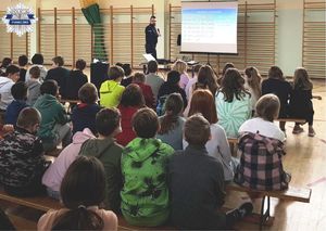 Policjant stojący przy tablicy multimedialnej, dzieci siedzące na ławkach. Na tablicy wyświetlana jest prezentacja dotycząca znaków drogowych