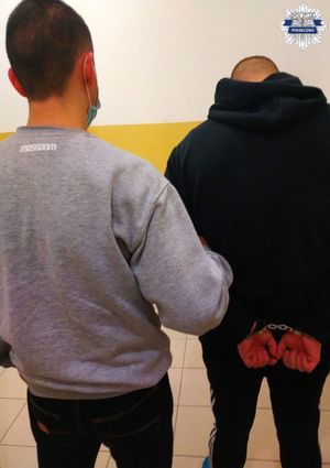 Zatrzymany mężczyzna wraz z policjantem. Mężczyzna ma założone kajdanki na rękach, stoją tyłem do aparatu