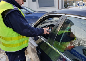 Policjant wydziału ruchu drogowego sprawdza stan trzeźwości kierującego pojazdem, kierujący ma uchylone okno w samochodzie. Widoczny jest tylko policjant