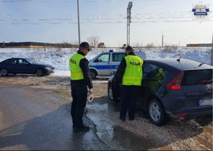 Policjant przy samochodzie, kierujący ma uchyloną szybę w swoim pojeździe, dokonywana jest kontrola stanu trzeźwości za pomocą urządzenia