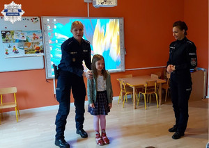 Policjantki stoją na środku sali razem z dziewczynką która opowiada innym dzieciom jak należy prawidłowo przechodzić przez jezdnię