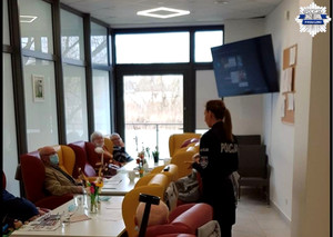 Policjantka stoi, seniorzy siedzą przy stolikach, prowadzona jest rozmowa na temat oszustw