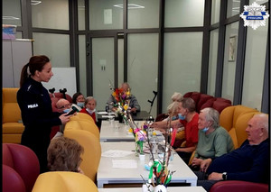 Policjantka stoi, seniorzy siedzą w fotelach przy stolikach, trwa rozmowa na temat oszustw