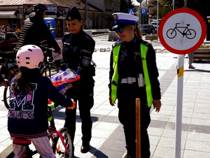 Policjantka nagradza dziewczynkę na rowerze prezentami za prawidłowe pokonanie toru przeszkód. Dziewczynka jest na rowerze w kasku