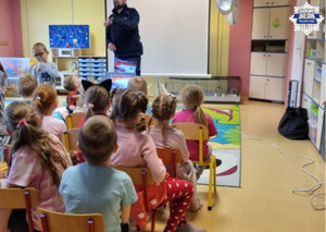 Dzieci siedzą na krzesłach, policjant stoi przed nimi i prowadzi prelekcję na temat bezpieczeństwa