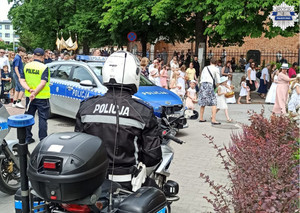 Policjant siedzący na motocyklu, drugi policjant stoi, w tle mieszkańcy powiatu uczestniczą w procesji Bożego Ciała