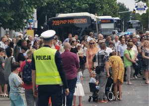 Policjant stoi, w tle autobus miejski z napisem Piaseczno oraz mieszkańcy uczestniczący w obchodach święta Bożego Ciała
