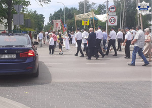 W tle nieoznakowany radiowóz z wyświetlanym napisem &quot;Policja&quot; oraz mieszkańcy biorący udział w procesji
