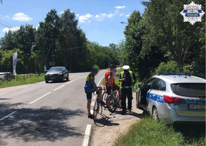 Policjant dokonujący kontroli rowerzystów, radiowoz stoi zaparkowany na poboczu, rowerzysci stoją obok radiowozu, policjant obok