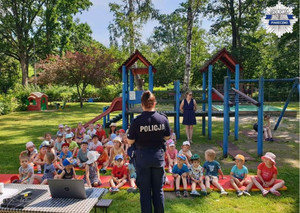 Policjantka stoi przodem do dzieci, przedszkolaki siedzą na materacach rozłożonych na trawie, trwa pogadanka