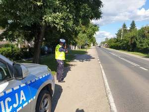 Policjant na poboczu drogi dokonuje pomiaru prędkości. Obok jest zaparkowany oznakowany radiowóz.