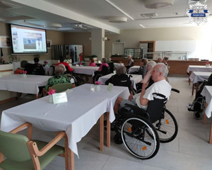 Pomieszczenie w budynku w którym są osoby starsze siedzące na wózkach inwalidzkich. W pomieszczeniu jest telebim, na którym są wyświetlane slajdy.