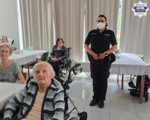 Pomieszczenie budynku, w którym na wózku inwalidzkim siedzi starsza kobieta a za nią jest stojąca policjantka w mundurze mająca zasłoniętą maseczką usta i nos.