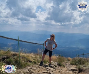 Kobieta w stroju sportowym stoi na szczycie góry w tle widać inne szczyty gór