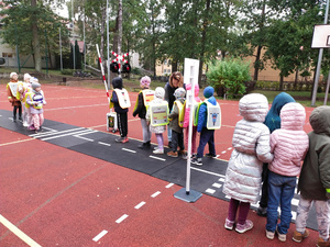 Na zdjęciu są dzieci ubrane w kurtkach, które ćwiczą zasady bezpieczeństwa na drodze na mobilnym miasteczku ruchu drogowego, które jest rozłożone na boisku szkolnym.