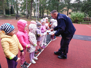 Na zdjęciu umundurowany policjant rozdaje dzieciom plany lekcji, które stoja w jednym rzędzie na boisku