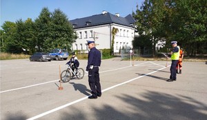 Umundurowany policjant z ruchu drogowego stoi a obok niego jedzie dziecko na rowerze i w kasku.