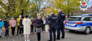 Policjanci stoją przy radiowozie, uczniowie stoją przed policjantami, spotkanie trwa na parkingu