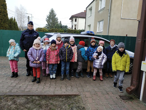 Policjantka ubrana w mundur stoi razem z przedszkolakami ubranymi w kurtki zimowe na tle radiowozu.