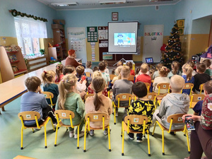Dzieci w przedszkolu siedzą na dywanie i ogladają bajkę wyświetlana na dużym wyświetlaczu.