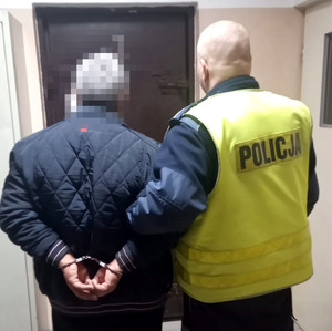 umundurowany policjant wydziału ruchu drogowego wraz z zatrzymanym mężczyzną, stoją tyłem, mężczyzna ma założone kajdanki na ręce trzymane z tyłu, zdjęcie wykonane jest w jednostce Policji
