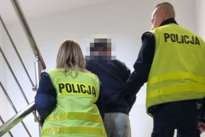 Mężczyzna z założonymi na ręce kajdankami, prowadzony przez umundurowanych policjantów