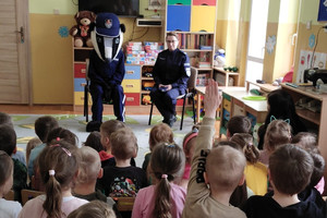 zdjęcie wykonane w sali przedszkolnej, dzieci siedzą na krzesełkach, jedno z nich ma uniesioną rękę. Przed dziećmi siedzi umundurowana policjantka oraz maskotka Komendy Stołecznej Policji - sierżant Borsuk