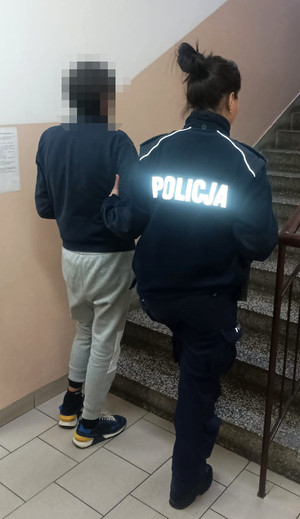 Umundurowana policjanta wraz z zatrzymanym mężczyzną. Zdjęcie zrobione jest przy schodach w jednostce Policji. Obydwoje odwróceni są tyłem.