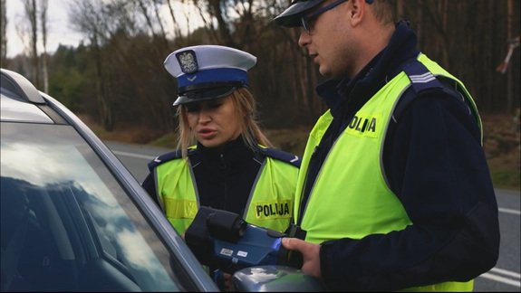 umundurowany policjant oraz policjantka wydziału ruchu drogowego stoją przy kontrolowanym samochodzie, policjant trzyma w ręku urządzenie służące do dokonywania pomiarów prędkości pokazując kierującej przekroczenie prędkości