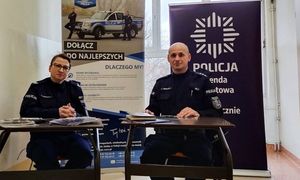 umundurowani policjanci siedzący przy stanowisku policyjny, w tle są banery informacyjne