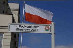powiewająca flaga Polski, tabliczka z nazwą ulicy &quot;podkomisarza Mirosława Żaka&quot;