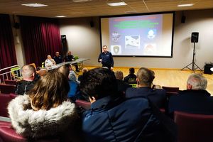 na sali widowiskowej siedzą mieszkańcy, przed nimi stoi umundurowany policjant który prowadzi spotkanie