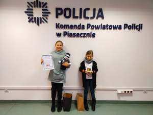 Na zdjęciu stoją dwie dziewczynki, które w rekach trzymają otrzymane nagrody. Na ścianie widać napis: Komenda Powiatowa Policji w Piasecznie